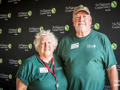 A man and woman wear green volunteer shirts and pose for a photo in front of a black TNC step-and-repeat banner.