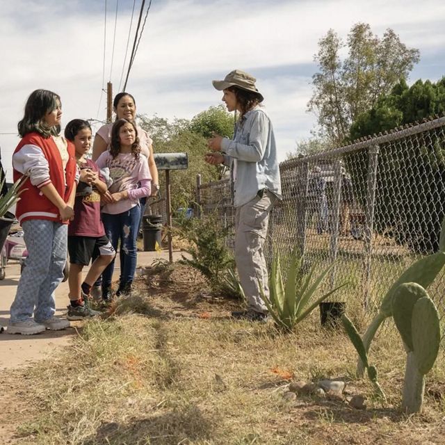 A woman standing by a fence talks to a group of three kids, one woman, and a man holding a potted plant.