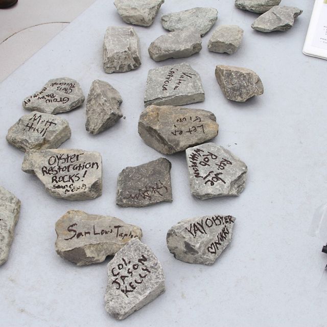 Small pieces of granite rock sit spread out on a table. Each piece of rock is signed with a message from people participating in an oyster reef dedication ceremony.