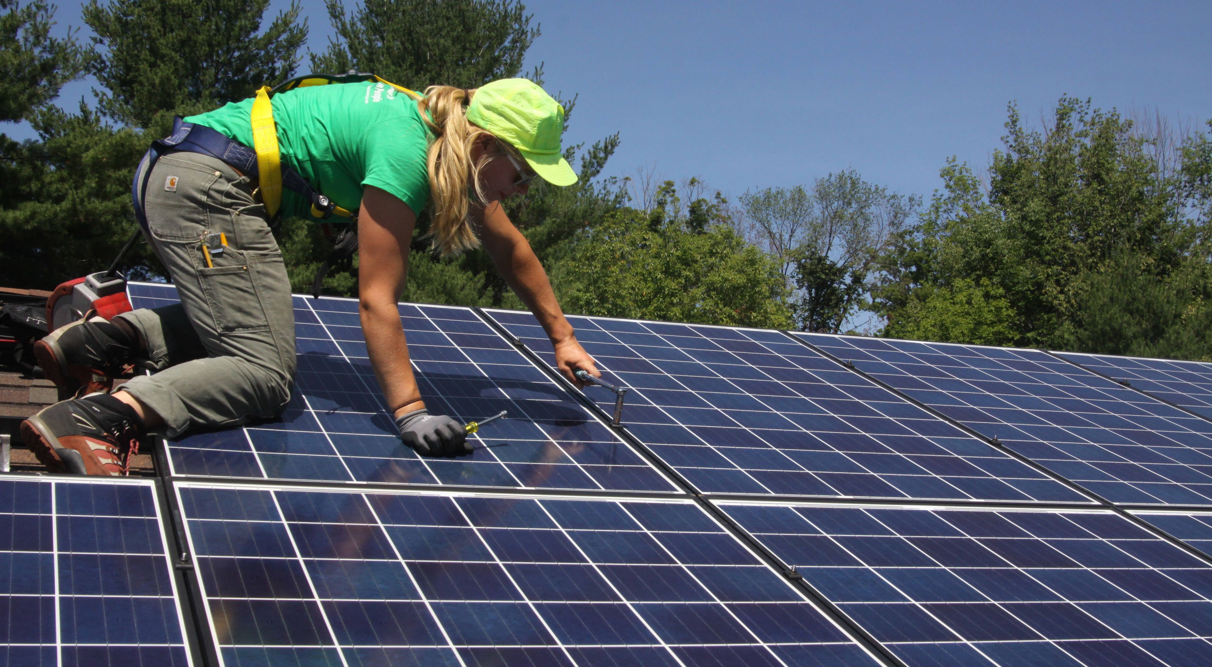 Solar panel installer on lefthand side of view crouching on top of a solar panel while screwing hardware into the panel. Green trees against a blue sky are in the background. 