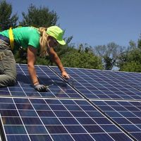 A solar energy installer working on a solar panel with green trees in the background.