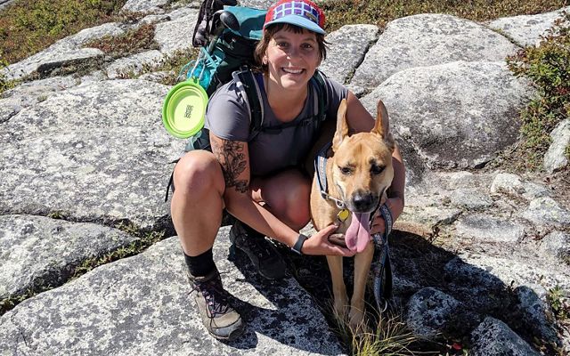 Una mujer con equipo de senderismo arrodillada en la cima de una montaña rocosa junto a su perro marrón.