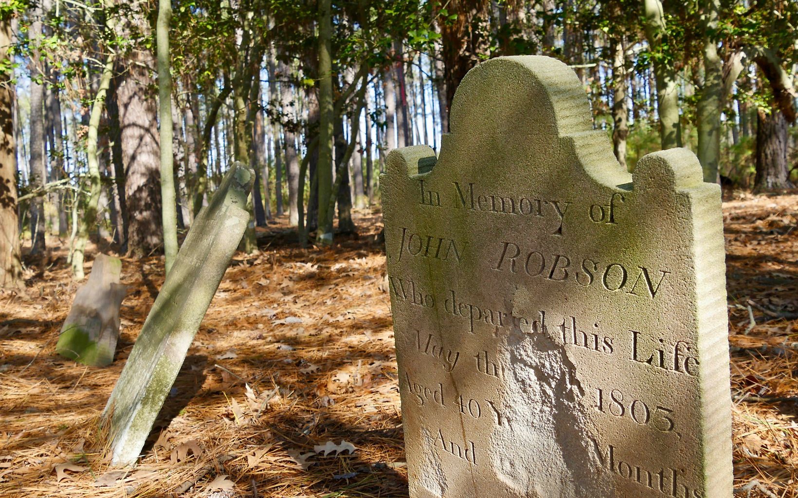 A row of headstones. The nearest marker reads, "In memory of John Robson Who departed this Life May 1805 Aged 40 years." The center of the marker has eroded away. The stone next to it is tiled back.