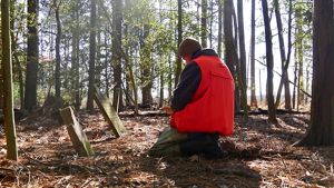 TNC Project Manager Joe Fehrer kneels in front of two tiled headstones. A man wearing a watch cap and blaze orange vest records data in a historic cemetery in the middle of a pine clearing.
