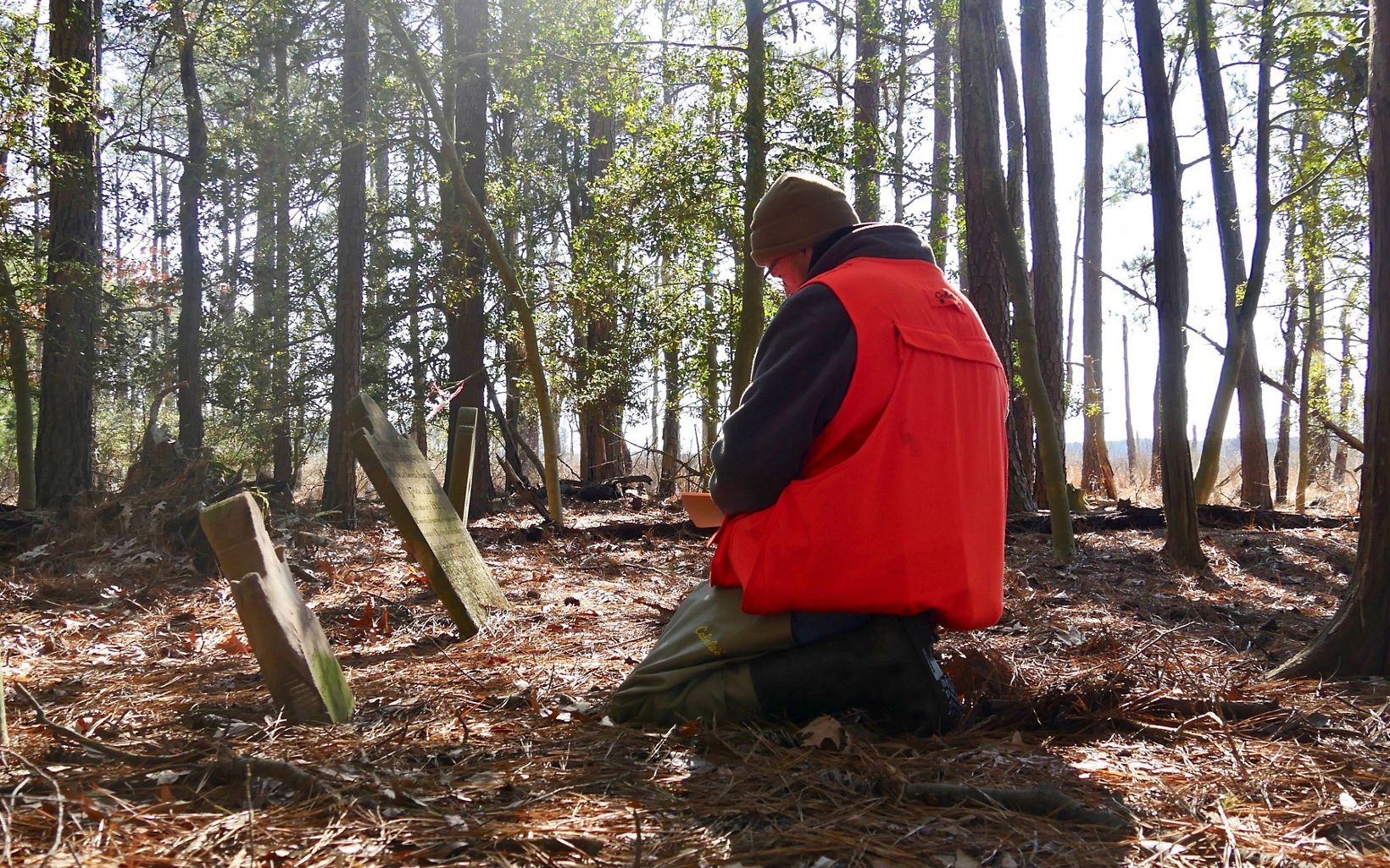 TNC Project Manager Joe Fehrer kneels in front of two tiled headstones. A man wearing a watch cap and blaze orange vest records data in an historic cemetery in the middle of a pine clearing.