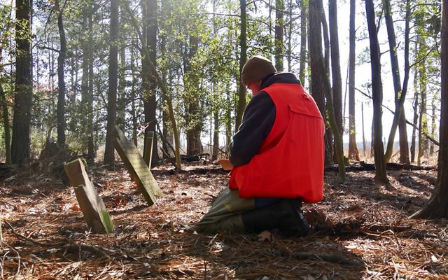 A man kneels on the ground in front of two weathered gravestones, recording the information on the markers.