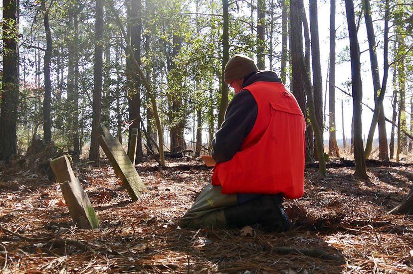 A man kneels on the ground in front of two weathered gravestones, recording the information on the markers.