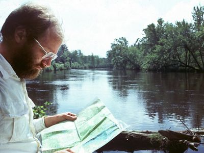 In the foreground, a bearded man looks at a map in front of a lake. 