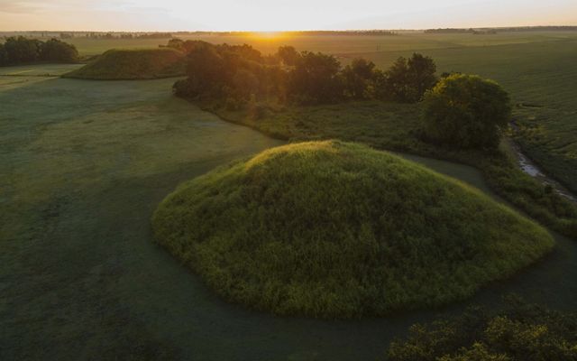 A grass covered mound rises above a flat field in Mississippi.