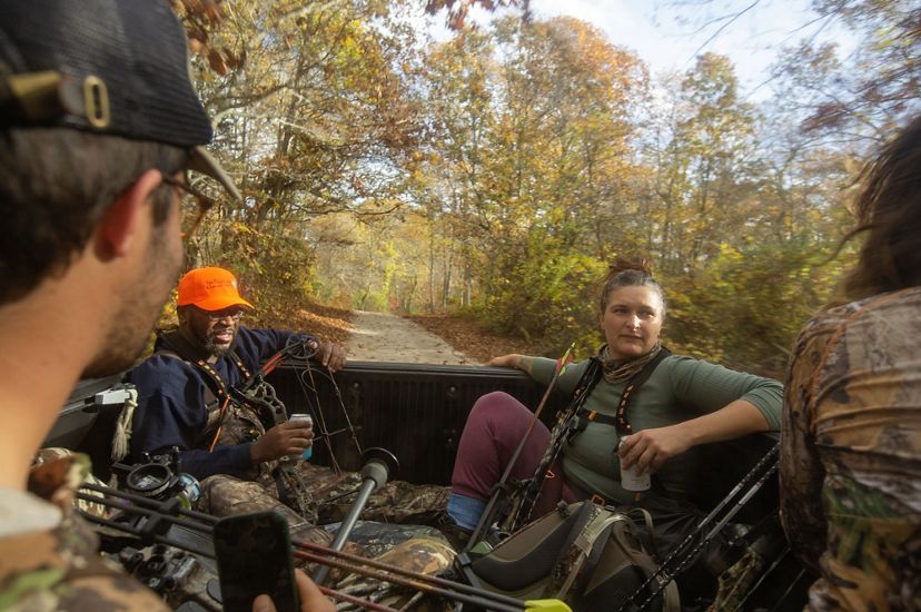 Several hunters ride in the bed of a truck through the woods, holding their gear.