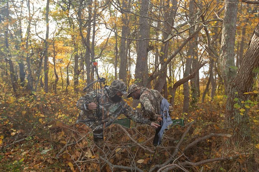 Amid a lot of autumn-colored brush in the woods, one man in camo helps another man in camo put on or take off a jacket.