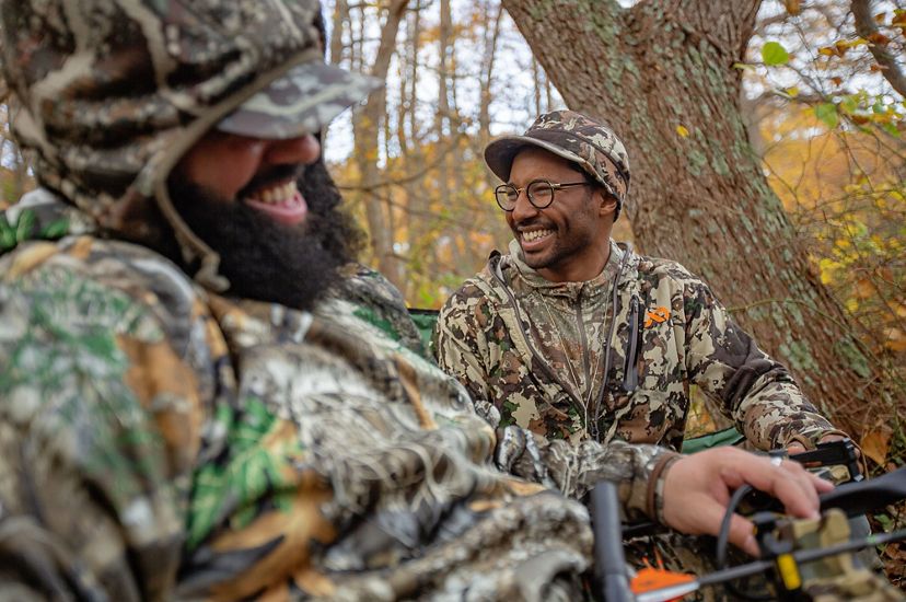 A close-up image of 2 men of color wearing camo and laughing together, sitting in the woods.