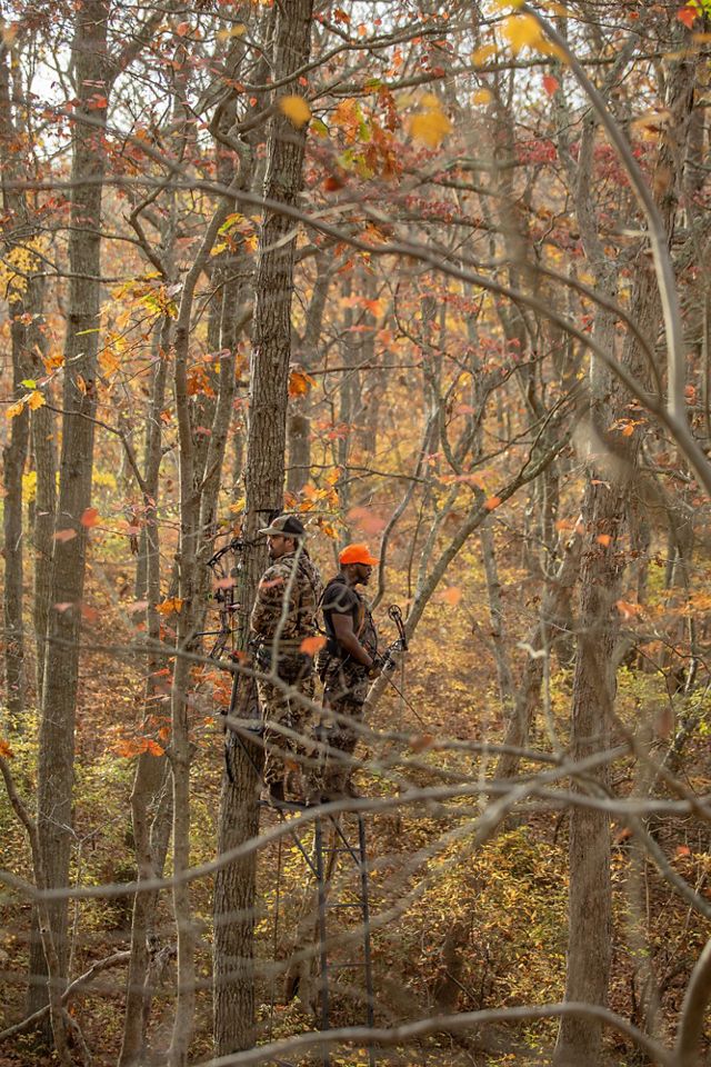 Two men wearing camouflage stand on a platform in the woods surrounded by autumn color. They hold crossbows.