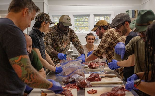 8 adults gather around a long metal table, each with cutting boards, meat and ziploc bags. 