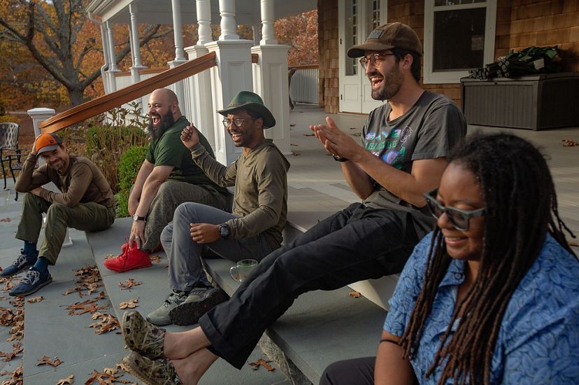 5 adults sit on the steps of the front porch of a building, all smiling, laughing or clapping. Fall color can be seen on the trees in the background.