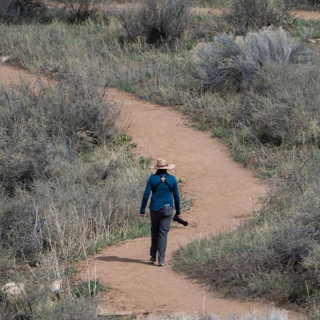 A person wearing a broad-brimmed hat and carrying a camera with a long lens walks away from the camera along a winding dirt path amid a scrubby landscape.