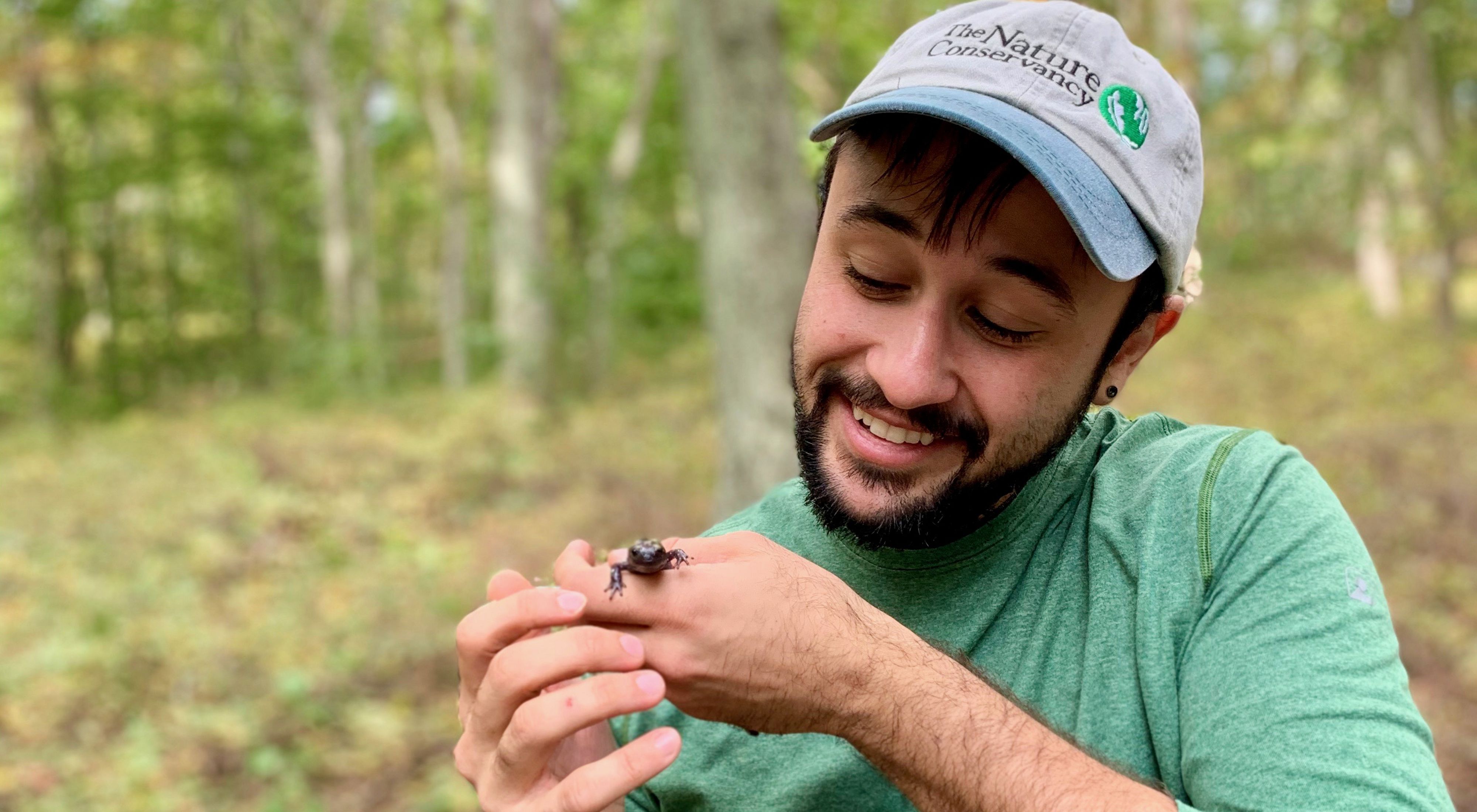 A person wearing a "The Nature Conservancy" baseball cap on the right smiling and looking at a salamander they are holding.