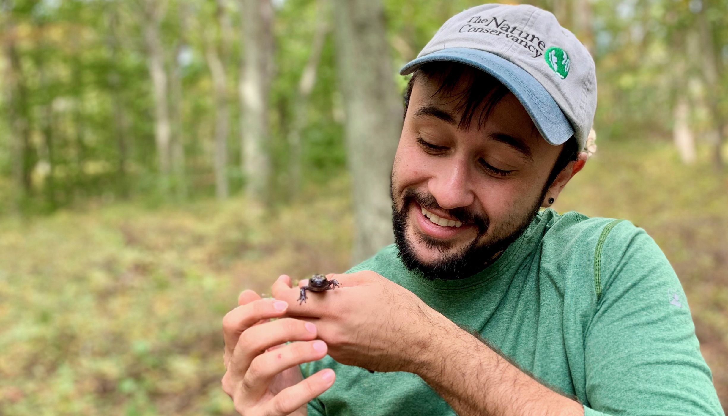 La imagen de una persona que tiene puesta una gorra de béisbol que dice "The Nature Conservancy", sonríe y está mirando una salamandra que tiene en la mano.