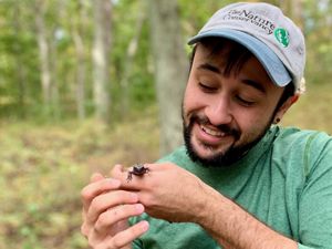 La imagen de una persona que tiene puesta una gorra de béisbol que dice "The Nature Conservancy", sonríe y está mirando una salamandra que tiene en la mano.