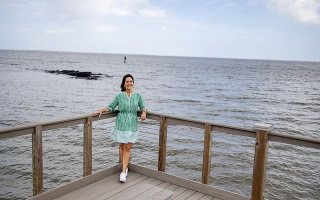 Jennifer VanVrancken, in a striped green dress and white tennis shoes, stands on the corner of a boardwalk over the water. In the background, a pile of rocks pokes above the rippling water. 