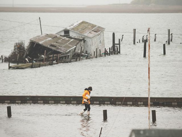 A woman ducks her head into the wind and she walks through ankle deep flood water. Behind her a building is partially collapsed in the water.