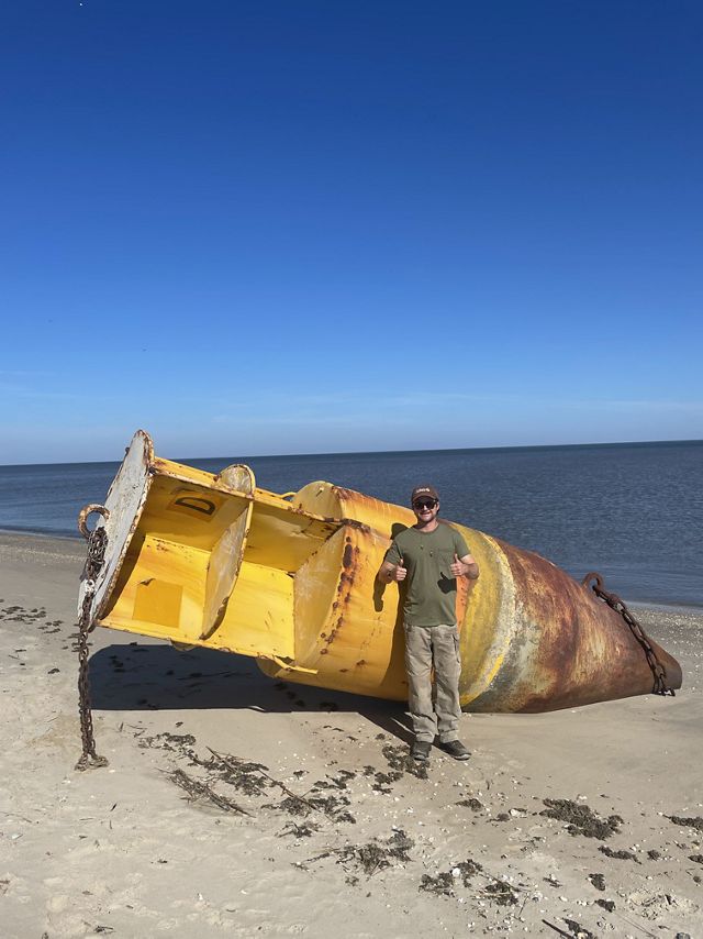 A photo of Russel on the beach in front of a large metal object that has washed up to shore.