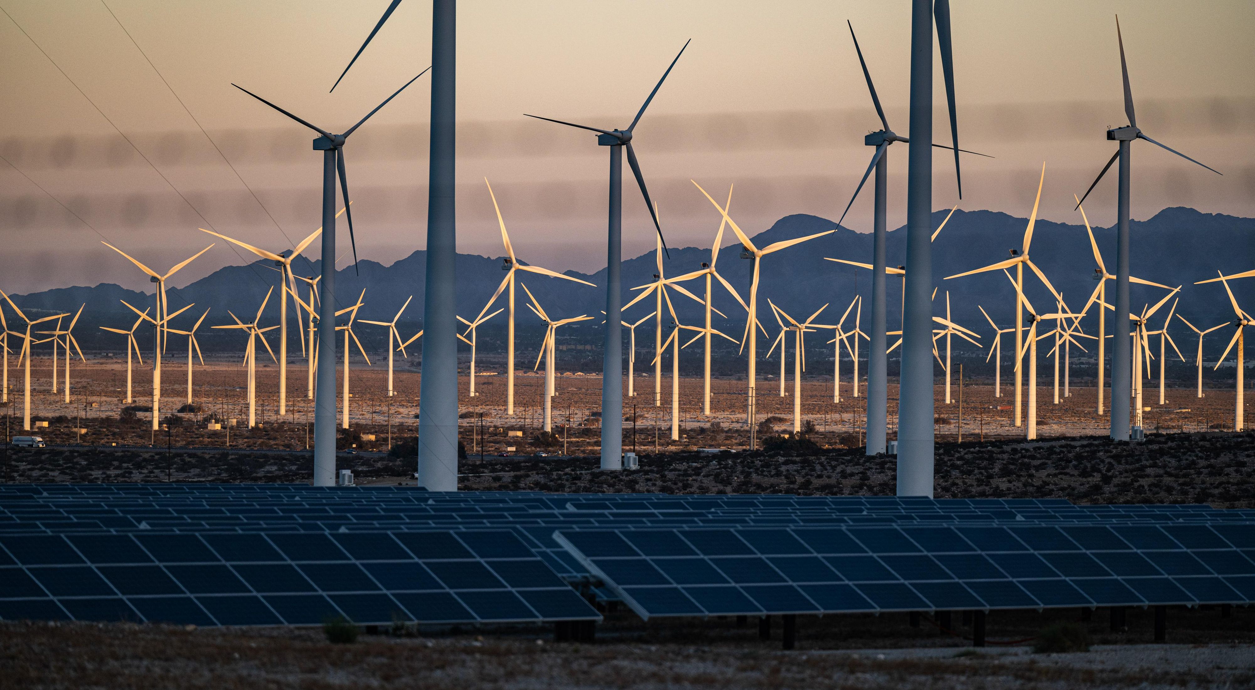 Photo during sunset of dozens of wind turbines towering over solar panels in the U.S. desert.