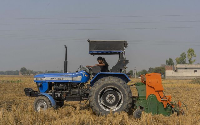 Amandeep drives through a dry rice field with a blue tractor with an orange attachment in back known as the super seeder. 