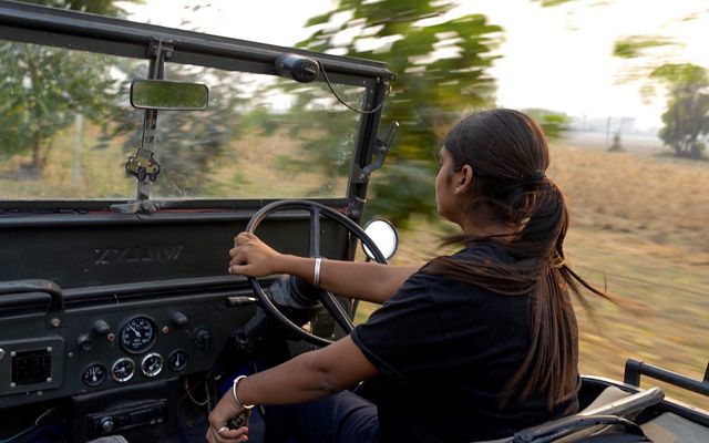 A view from the passenger side of an open jeep as Amandeep Kaur drives through a farming region, the landscape flying in motion around her.