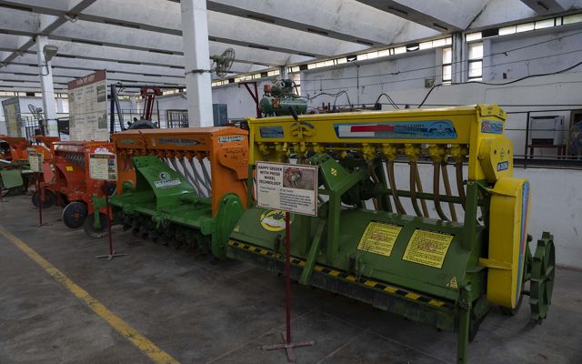 In a long university building, 3 large seeder machines of different colors sit next to each other, full of rotors and hoses.