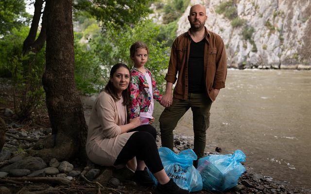 A family stand on the banks of a river with bags of items for a picnic.