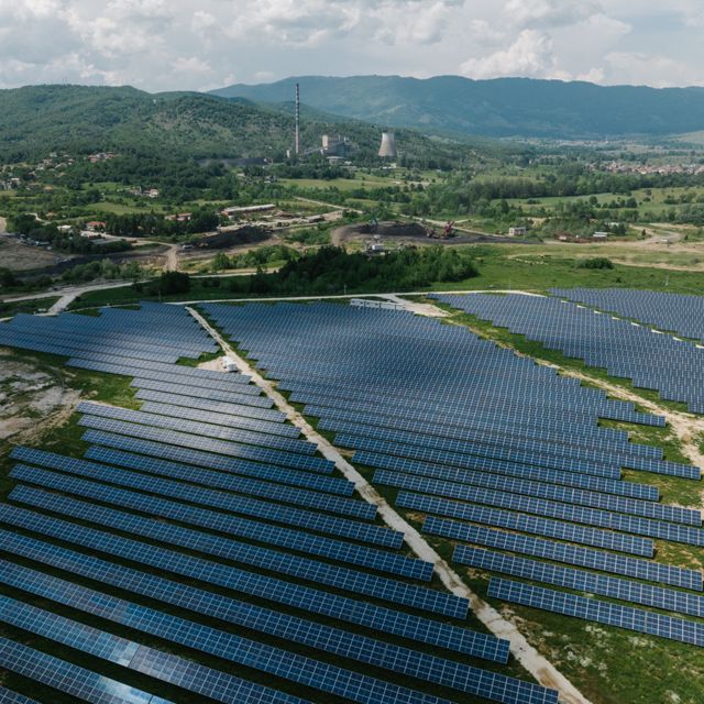 Tight lines of solar panels fill a grassy field with coal mining infrastructure in the background.