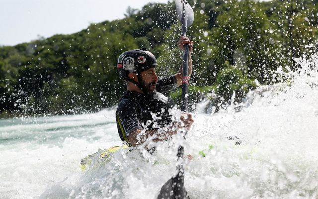 A kayaker wearing a helmet navigates through splashes and whitewater rapids on a sunny day.