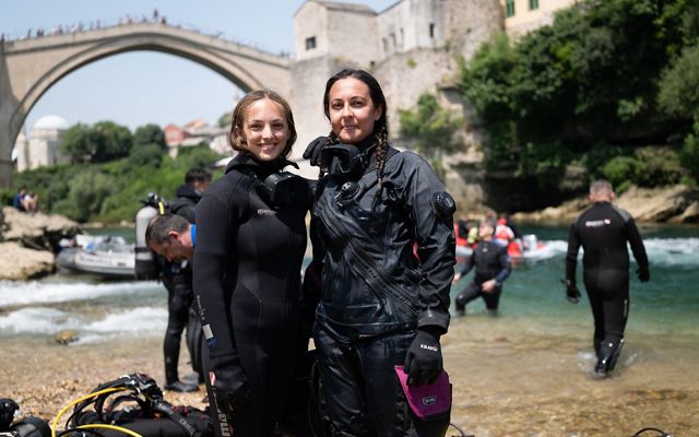 Two divers in wet suits smile and pose on a river bank with an old arched stone bridge in the background.