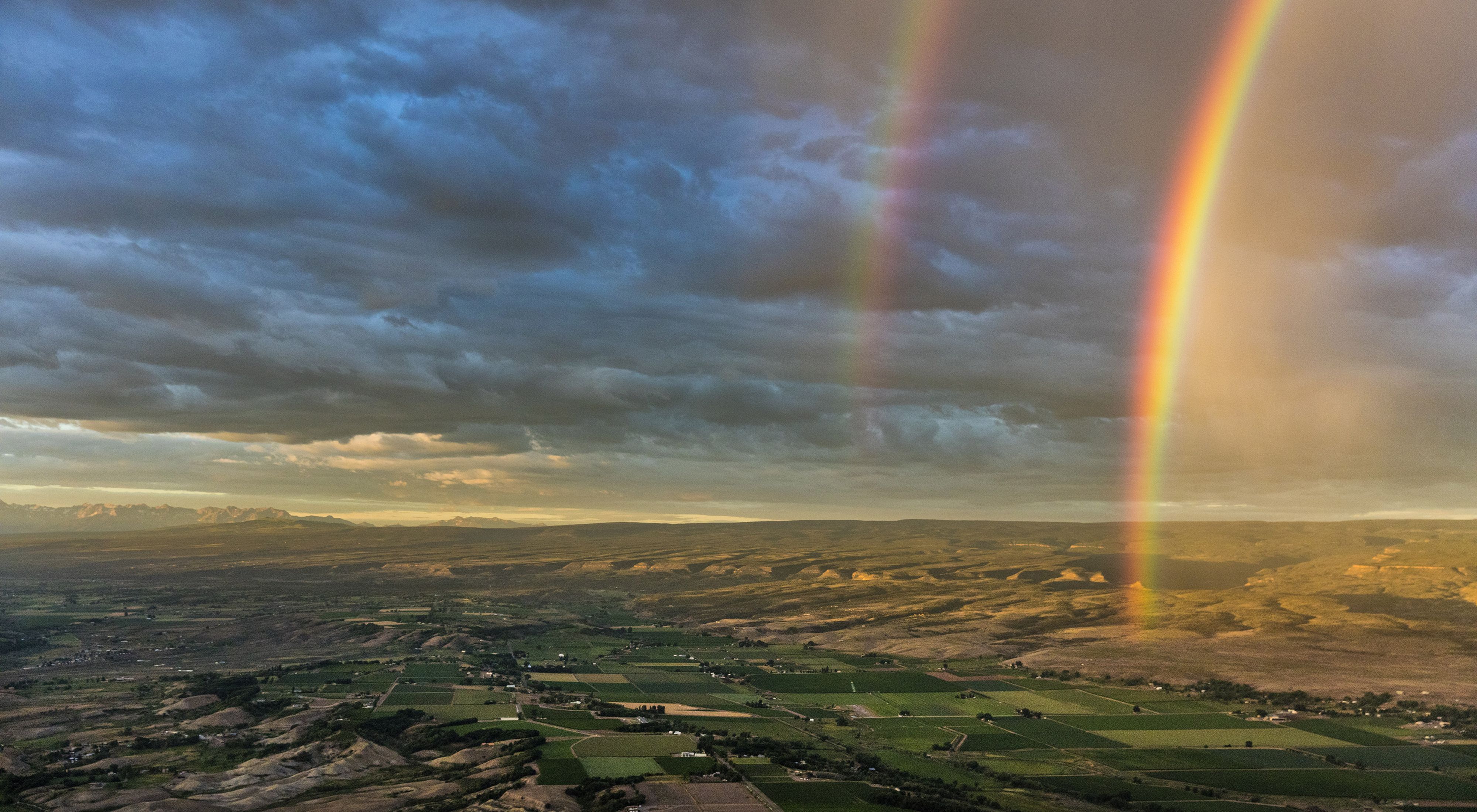 Aerial view of a vast landscape of agricultural field, homes, hills, and mountains with a double rainbow appearing over the fields.