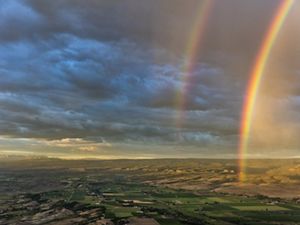 Aerial view of a vast landscape of agricultural field, homes, hills, and mountains with a double rainbow appearing over the fields.