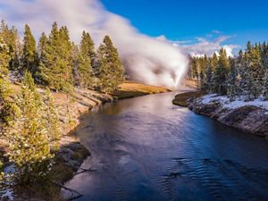A river flows up to a steaming geyser, lined on both sides with a rocky shore and evergreen trees, with snow in the shadows.