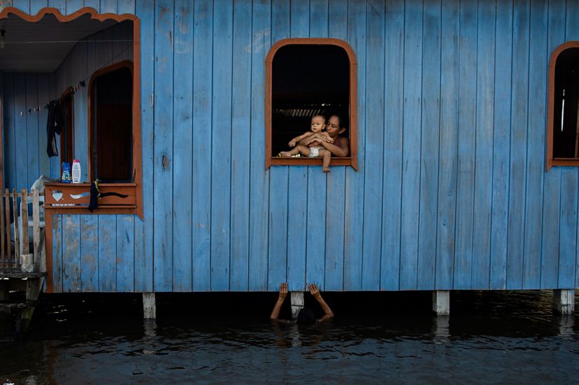 Una mujer sentada en la ventana de una casa azul, sosteniendo a un bebé en brazos. Debajo de ella, un niño la mira desde las aguas profundas que fluyen debajo de la casa.