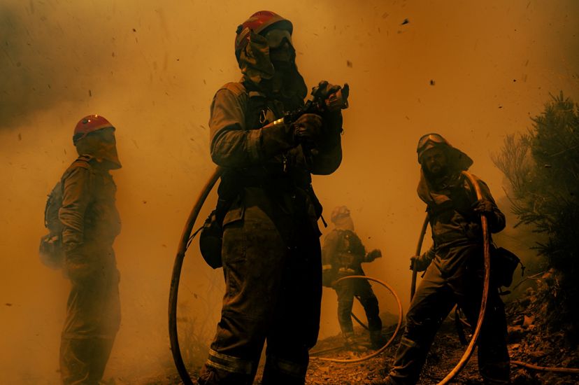Cuatro bomberos sostienen una manguera de agua y se encuentran al borde de un bosque. El aire es anaranjado y lleno de humo.