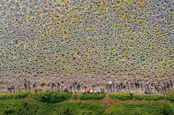 A group of people stand at the edge of a large, open sandy area. Large holes dot the surface, filled with green and orange water.