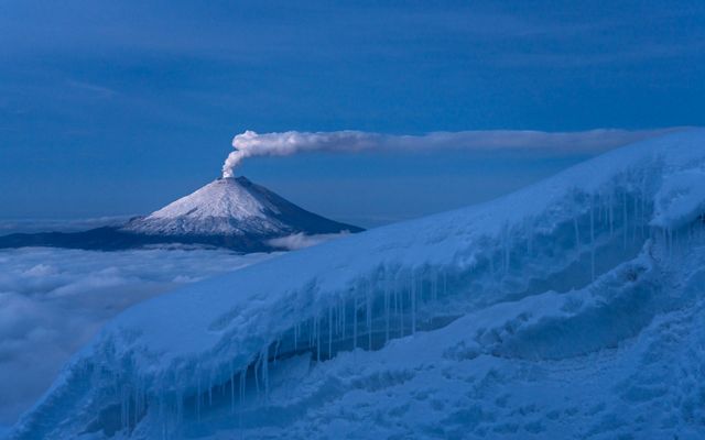 A column of steam rises from the snow capped dome of a volcano. Clouds fill the valley between the volcano and the the snow covered glacier in the foreground.