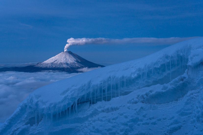 A column of steam rises from the snow capped dome of a volcano. Clouds fill the valley between the volcano and the the snow covered glacier in the foreground.