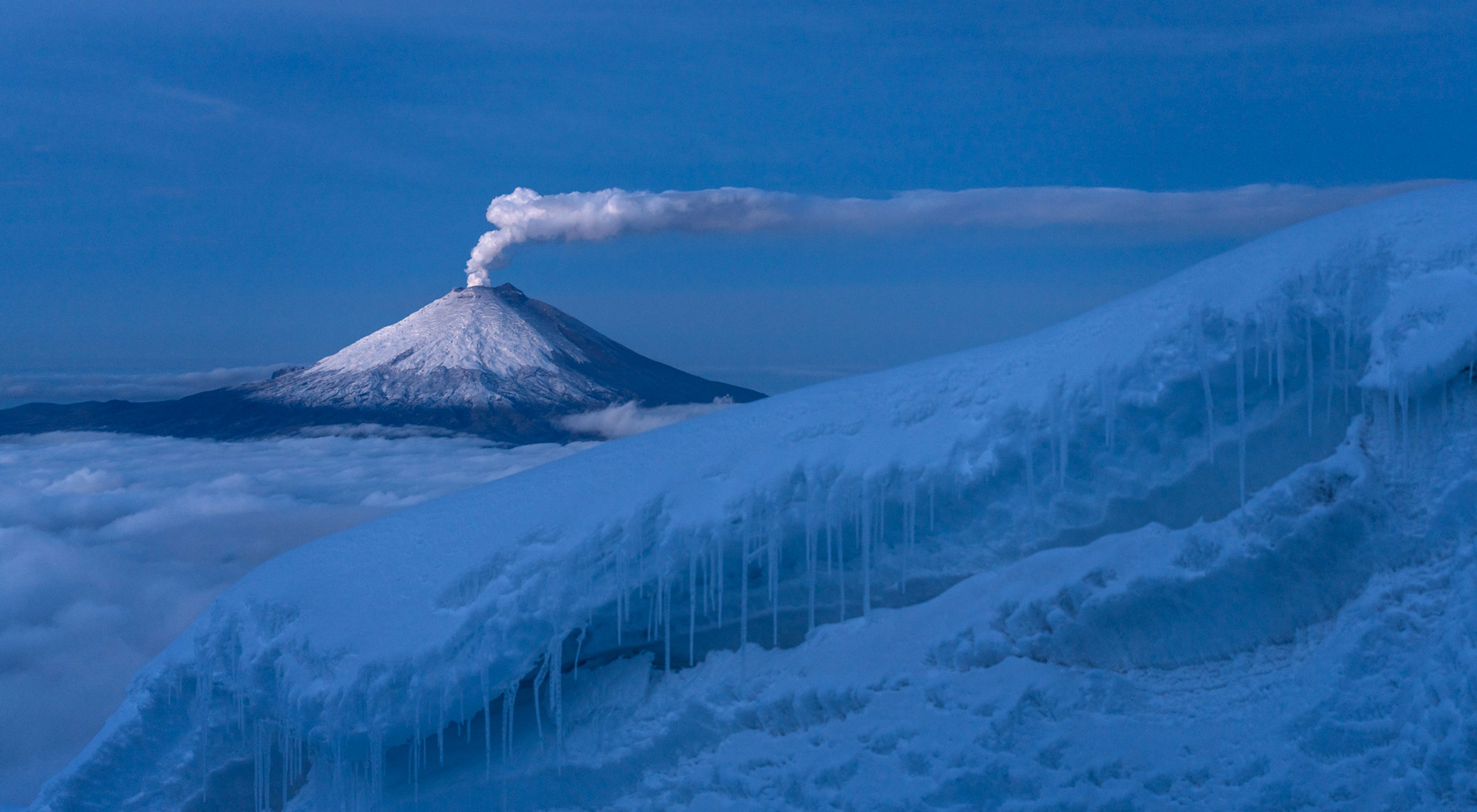 Cotopaxi volcano spews a long column of steam from a distance over a snowy landscape