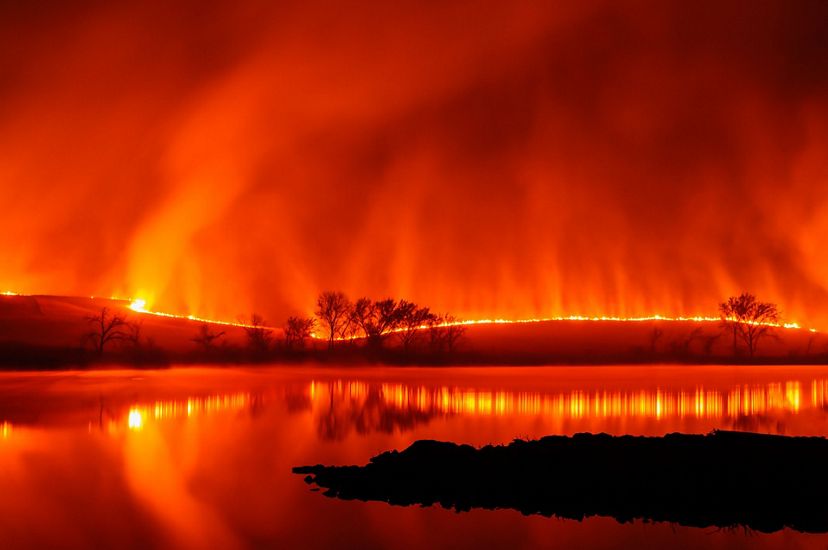 A glowing line of fire edges the horizon. Plumes of smoke rise in the orange tinged sky. The fire line is blurred and reflected in the surface of a lake.