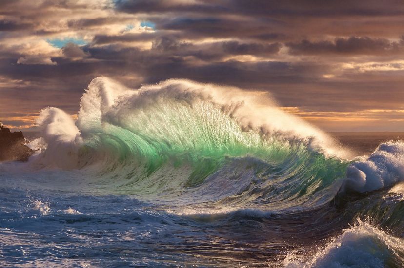 A massive wave crashes on the shore, creating a wide curtain of water. The base of the wave is pale green. The white foam spray blends into the low hanging storm clouds.