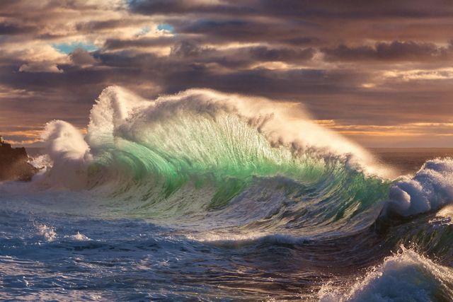 A massive wave crashes on the shore, creating a wide curtain of water. The base of the wave is pale green. The white foam spray blends into the low hanging storm clouds.