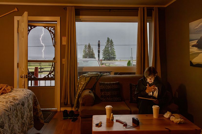 A woman sits on a sofa, her face softly illuminated by the glow of her tablet screen. A thick bolt of lightening is visible through the open door, splitting the gray sky.