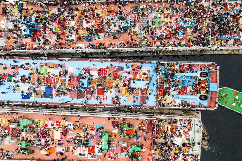 People and blankets create a brightly colored patchwork on the roof decks of three long boats. A smaller green decked boat floats at an angle behind the larger vessel in the center.