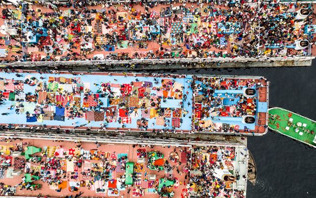 Personas y mantas crean un mosaico de colores brillantes encima de tres largos barcos.