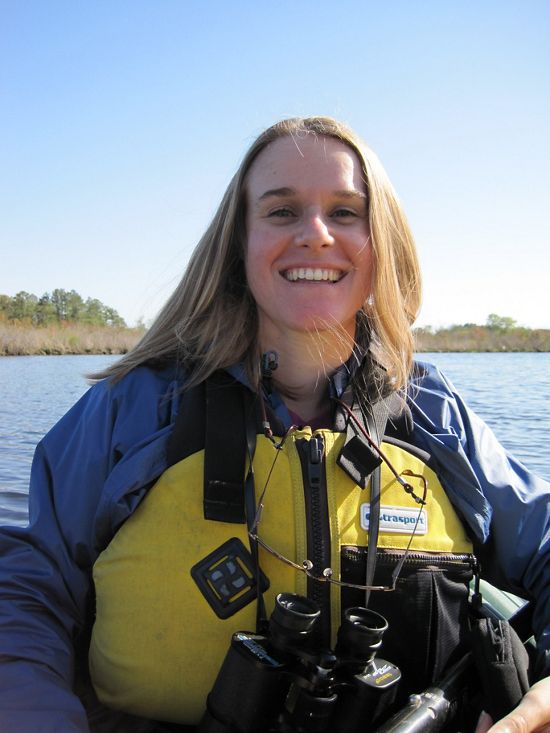 Tamara Gagnolet sits on a kayak in the water wearing a yellow life vest and binoculars. 